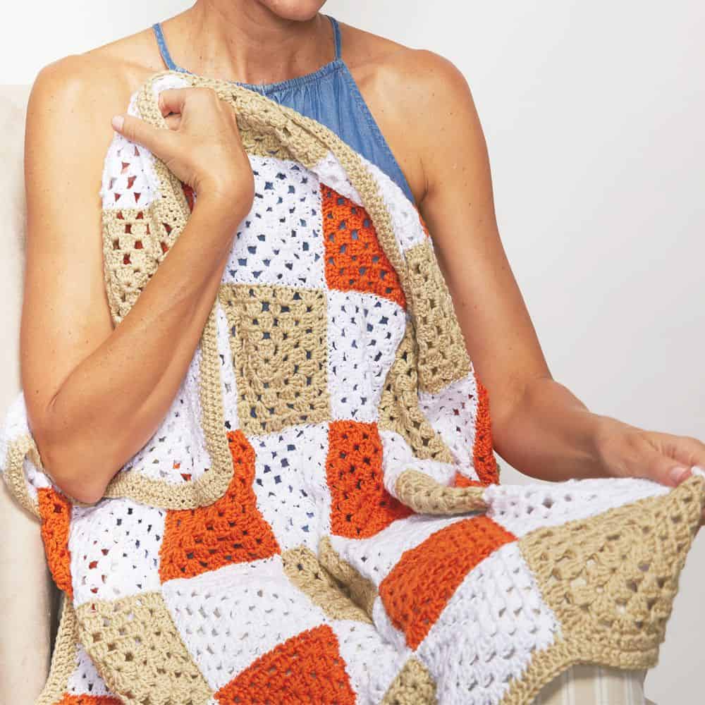 a person holding a finished granny square crochet blanket in white, beige and orange